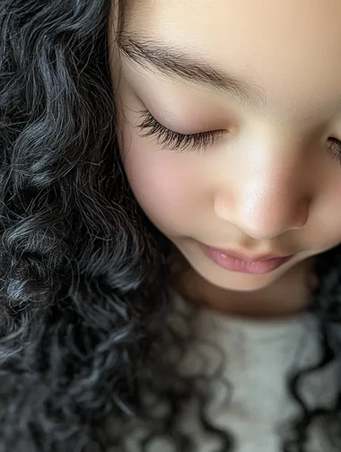 Realistic photos taken using an iPhone camera. A close-up portrait of a child with long, curly black hair and exceptionally long eyelashes. The child's face is positioned slightly below the center of the frame, with their eyes gently lowered. Soft, warm natural light falls from the upper left, creating delicate highlights on the child's smooth skin and casting subtle shadows that enhance facial features. The background is a blurred, light-colored interior setting, maintaining a shallow depth of field to keep focus on the subject. The overall atmosphere is cozy and serene, with a warm color palette dominating the image. The camera angle is slightly above the child, looking down to capture the full length of the eyelashes. The lighting is gentle and diffused, emphasizing the texture of the hair and skin without harsh contrasts. The child's expression is calm and peaceful, contributing to the and tender mood of the portrait. Realistic photos taken using an iPhone camera.