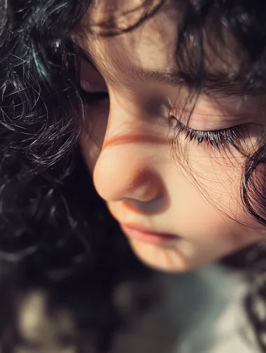Realistic photos taken using an iPhone camera. A close-up portrait of a child with long, curly black hair and exceptionally long eyelashes. The child's face is positioned slightly below the center of the frame, with their eyes gently lowered. Soft, warm natural light falls from the upper left, creating delicate highlights on the child's smooth skin and casting subtle shadows that enhance facial features. The background is a blurred, light-colored interior setting, maintaining a shallow depth of field to keep focus on the subject. The overall atmosphere is cozy and serene, with a warm color palette dominating the image. The camera angle is slightly above the child, looking down to capture the full length of the eyelashes. The lighting is gentle and diffused, emphasizing the texture of the hair and skin without harsh contrasts. The child's expression is calm and peaceful, contributing to the and tender mood of the portrait. Realistic photos taken using an iPhone camera.