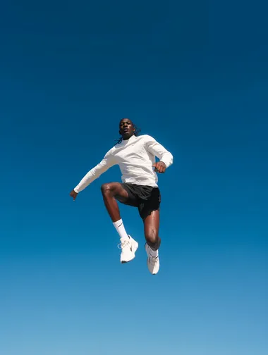 A dynamic, front-angle shot of a runner mid-jump against a clear blue sky. The runner is wearing a white long-sleeve top, black shorts, white socks, and white running shoes. The pose is casual and energetic, with the runner's knees bent and arms slightly extended backward as they jump, creating a sense of power and movement. Clean, minimal background with no distractions, capturing a modern, motivational sports aesthetic ideal for a Nike running advertisement.