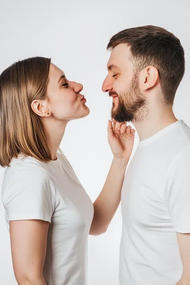a photograph of side view of white female around 35 years blowing kisses in the air toward right cheek of white male. white male is looking straight at us smiling at us. both wearing white tees in white background.