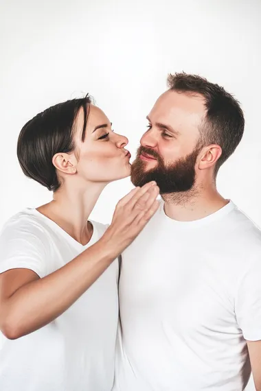 a photograph of side view of white female around 35 years blowing kisses in the air toward right cheek of white male. white male is looking straight at us smiling at us. both wearing white tees in white background.