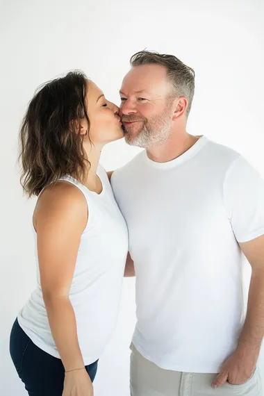 a photograph of side view of white female around 35 years blowing kisses in the air toward right cheek of white male. white male is looking straight at us smiling at us. both wearing white tees in white background.