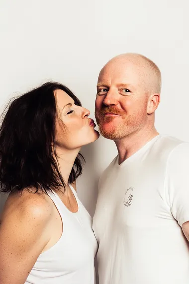 a photograph of side view of white female around 35 years blowing kisses in the air toward right cheek of white male. white male is looking straight at us smiling at us. both wearing white tees in white background.