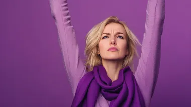 Profile view of an angry blonde woman with a purple scarf around her neck protesting raising her arms on purple background with plenty of copy space, international women's day