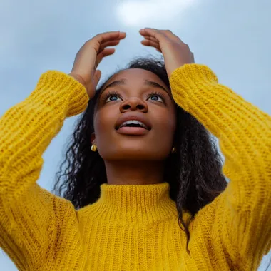 african american looking shocked but happy looking up in the sky, holding her hands to her head, wearing a long sleeved yellow sweater