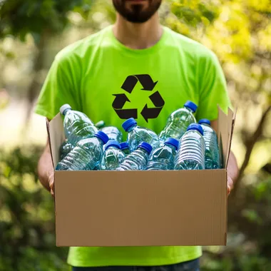 A realistic image depicting a person holding a box filled with plastic water bottles, demonstrating recycling efforts. The individual is wearing a bright green shirt with a recycling symbol. The background is a natural outdoor setting with trees and greenery, emphasizing environmental awareness. The color scheme should feature green tones, white for the box, and blue shades from the water bottles.