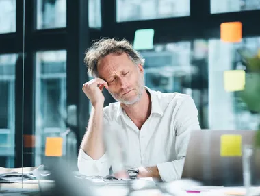 A middle-aged man sits at a desk in a modern office, appearing stressed or exhausted, with his head resting in one hand and eyes closed. The workspace includes a laptop, some papers, and large windows in the background that let in natural light. Sticky notes are visible on the glass, suggesting a work environment with ongoing tasks or deadlines. He is wearing a white shirt, which can be changed to a soft grey-blue or muted olive tone to create a more visually balanced and emotionally resonant scene. This is a stock photo with ultra high detail, photo realism, and maximum skin realism. The image should convey hyperrealism through expressive facial details, natural lighting, and realistic skin texture. The shirt color must differ from the original, using a harmonious palette that fits the emotional tone of the image. The overall mood should remain authentic and relatable within a professional setting.  --raw