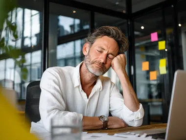 A middle-aged man sits at a desk in a modern office, appearing stressed or exhausted, with his head resting in one hand and eyes closed. The workspace includes a laptop, some papers, and large windows in the background that let in natural light. Sticky notes are visible on the glass, suggesting a work environment with ongoing tasks or deadlines. He is wearing a white shirt, which can be changed to a soft grey-blue or muted olive tone to create a more visually balanced and emotionally resonant scene. This is a stock photo with ultra high detail, photo realism, and maximum skin realism. The image should convey hyperrealism through expressive facial details, natural lighting, and realistic skin texture. The shirt color must differ from the original, using a harmonious palette that fits the emotional tone of the image. The overall mood should remain authentic and relatable within a professional setting.  --raw