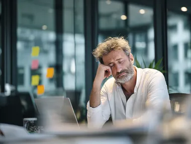 A middle-aged man sits at a desk in a modern office, appearing stressed or exhausted, with his head resting in one hand and eyes closed. The workspace includes a laptop, some papers, and large windows in the background that let in natural light. Sticky notes are visible on the glass, suggesting a work environment with ongoing tasks or deadlines. He is wearing a white shirt, which can be changed to a soft grey-blue or muted olive tone to create a more visually balanced and emotionally resonant scene. This is a stock photo with ultra high detail, photo realism, and maximum skin realism. The image should convey hyperrealism through expressive facial details, natural lighting, and realistic skin texture. The shirt color must differ from the original, using a harmonious palette that fits the emotional tone of the image. The overall mood should remain authentic and relatable within a professional setting.  --raw