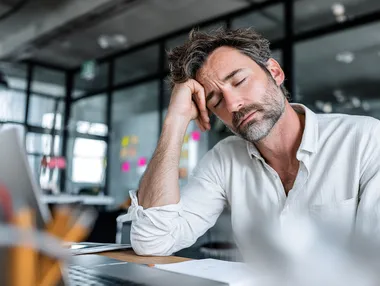 A middle-aged man sits at a desk in a modern office, appearing stressed or exhausted, with his head resting in one hand and eyes closed. The workspace includes a laptop, some papers, and large windows in the background that let in natural light. Sticky notes are visible on the glass, suggesting a work environment with ongoing tasks or deadlines. He is wearing a white shirt, which can be changed to a soft grey-blue or muted olive tone to create a more visually balanced and emotionally resonant scene. This is a stock photo with ultra high detail, photo realism, and maximum skin realism. The image should convey hyperrealism through expressive facial details, natural lighting, and realistic skin texture. The shirt color must differ from the original, using a harmonious palette that fits the emotional tone of the image. The overall mood should remain authentic and relatable within a professional setting.  --raw