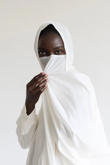 Create a high-resolution, full-body image of a white model standing and applying a fabric sheet mask to her face. The mask covers her entire face except for the eyes and lips, with no visible product or serum on the mask, showcasing a clean, smooth texture. The model faces the camera directly with a calm and relaxed expression. The studio lighting emphasizes the natural contours of her face, the fabric mask's texture, and the subtle details of her glowing skin