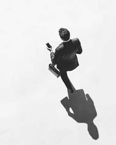 A black and white photograph of an overhead shot from above, a man in a suit holding his phone while walking with a briefcase, the shadow is cast on the ground by him. The background is white.