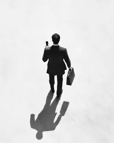 A black and white photograph of an overhead shot from above, a man in a suit holding his phone while walking with a briefcase, the shadow is cast on the ground by him. The background is white.