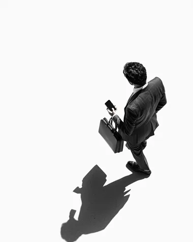 A black and white photograph of an overhead shot from above, a man in a suit holding his phone while walking with a briefcase, the shadow is cast on the ground by him. The background is white.