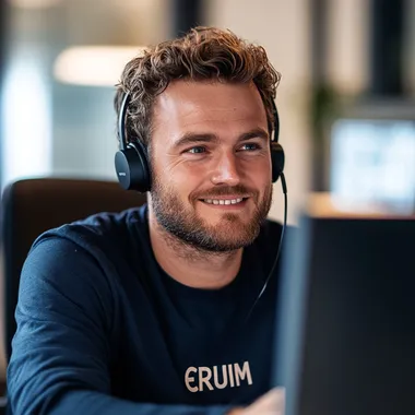 An ultra-realistic photograph captured with a Canon EOS 2000D, equipped with an 85mm lens at F 1.2 aperture setting, portraying an average-looking Dutch 30-year-old happy-looking man wearing a navy blue office attire with a navy blue t-shirt underneath that has CERUIM printed in bold white letters. He is wearing a call headset while looking motivated at his desktop screen, sitting at his desk while calling a client. The background is ever so slightly blurred. The image, shot in high resolution and a 9:16 aspect ratio, captures the subjects motivational and kind nature. ar 16:9 style raw