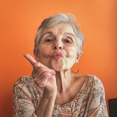A old woman 60 years with short grey hair is blowing a kiss to the camera in her home. She is dressed casually and wearing hoop earrings. The scene is captured from an angle that highlights both her face and upper body, background flat orange   --s 50