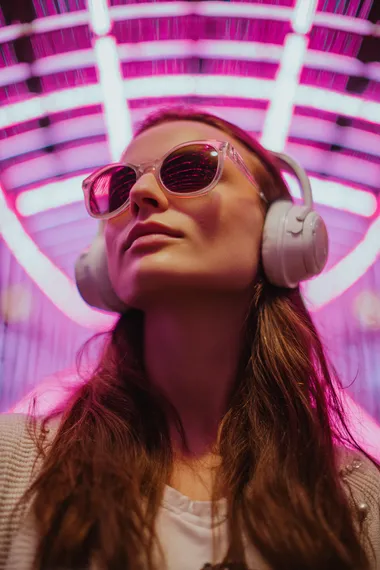 Flyer for a rave night featuring a young, fair-skinned woman, around 22 years old, with long brown hair, wearing sunglasses and white headphones, looking up. The background is a futuristic, neon-lit structure with a grid pattern, illuminated in vibrant pink.  --raw