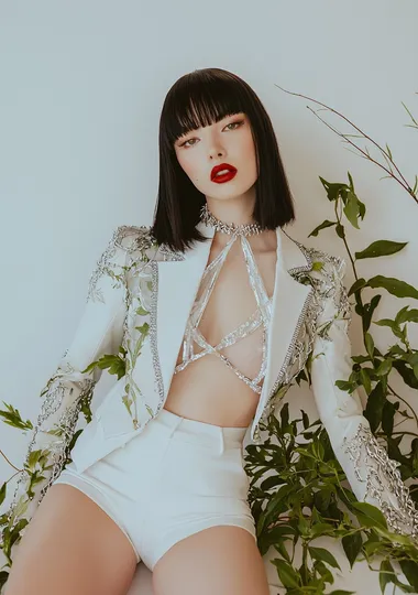 A beautiful model wearing a white skirt with a shirt and cropped jacket designed in the style of Alexander McQueen. The top has long sleeves with silver trim. She has shoulder length black straight hair with bangs and is wearing red lipstick, sitting on an all-white background. There is some greenery around her legs, and she is wearing a large chain necklace. This is a fashion photography editorial look for Vogue magazine