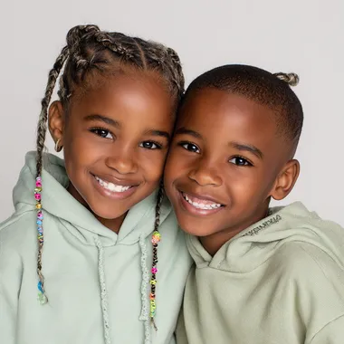 A close-up portrait of a young Black girl and boy with neatly braided hair adorned with colorful beads. they smile to the camera.they both have braid , the boy have STITCH BRAIDS, and the girl have TWIN BUN STITCH BRAIDS, and both wears a light pastel green hoodie . The background is minimal and light, giving a soft studio feel. background color is white. studio lighting, clean and high-resolution fashion photography style.