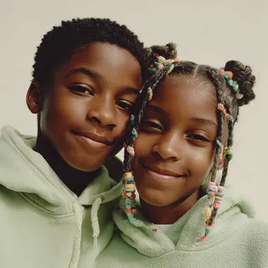 A close-up portrait of a young Black girl and boy with neatly braided hair adorned with colorful beads. they smile to the camera.they both have braid , the boy have STITCH BRAIDS, and the girl have TWIN BUN STITCH BRAIDS, and both wears a light pastel green hoodie . The background is minimal and light, giving a soft studio feel. background color is white. studio lighting, clean and high-resolution fashion photography style.