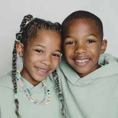 A close-up portrait of a young Black girl and boy with neatly braided hair adorned with colorful beads. they smile to the camera.they both have braid , the boy have STITCH BRAIDS, and the girl have TWIN BUN STITCH BRAIDS, and both wears a light pastel green hoodie . The background is minimal and light, giving a soft studio feel. background color is white. studio lighting, clean and high-resolution fashion photography style.