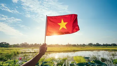 rough hands gently hold the Vietnamese flag in the clear blue sky,just have one five-pointed star in the middle of the flag ,straight flag, with gentle sunshine, below is a lotus field with leaves, buds, and blooming flowers, in the middle of the rice field is a river flowing through, 16K