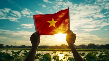 rough hands gently hold the Vietnamese flag in the clear blue sky,just have one five-pointed star in the middle of the flag ,straight flag, with gentle sunshine, below is a lotus field with leaves, buds, and blooming flowers, in the middle of the rice field is a river flowing through, 16K