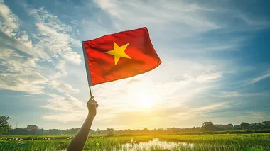 rough hands gently hold the Vietnamese flag in the clear blue sky,just have one five-pointed star in the middle of the flag ,straight flag, with gentle sunshine, below is a lotus field with leaves, buds, and blooming flowers, in the middle of the rice field is a river flowing through, 16K