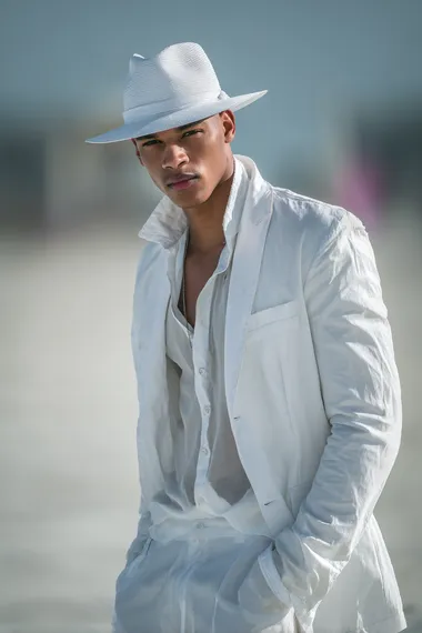 Create a stunning image of an LGBTQ beautiful male model wearing all white catwalk fashion clothing, in a beach environment, Nikon photo shoot quality