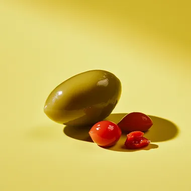 Straight on, very low eye level camera angle. Showcasing the side view of a half olive. add a group of three small chopped pieces red pimento next to the olive. Natural and soft lighting. Daylight bright setting. Placed on a yellow background. --s 50