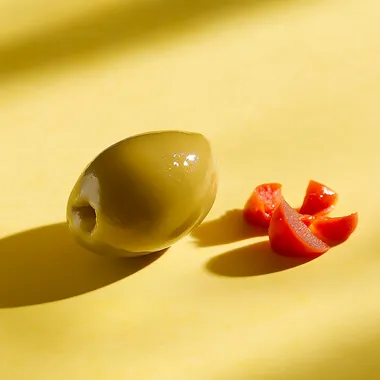 Straight on, very low eye level camera angle. Showcasing the side view of a half olive. add a group of three small chopped pieces red pimento next to the olive. Natural and soft lighting. Daylight bright setting. Placed on a yellow background. --s 50