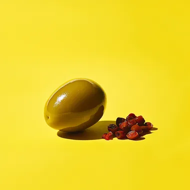Straight on, very low eye level camera angle. Showcasing the side view of a half olive. add a group of three small chopped pieces red pimento next to the olive. Natural and soft lighting. Daylight bright setting. Placed on a yellow background. --s 50