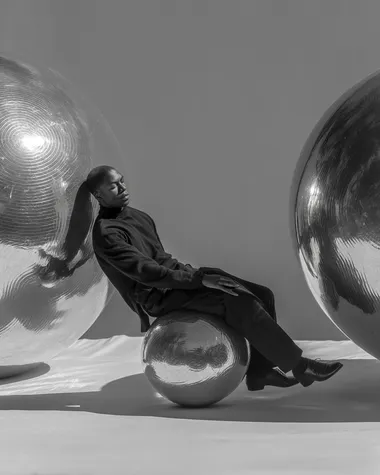 A surreal black-and-white image of a man lying horizontally between two large metallic spheres--one above him, one below. His body is gently pressed between them, as if suspended in stillness. He stretches his arms around the lower sphere, embracing it. The background is pure white, emphasizing the surreal, sculptural composition. The image feels dreamlike, introspective, and artistic, with strong contrast and soft, poetic lighting.   --s 350 --raw