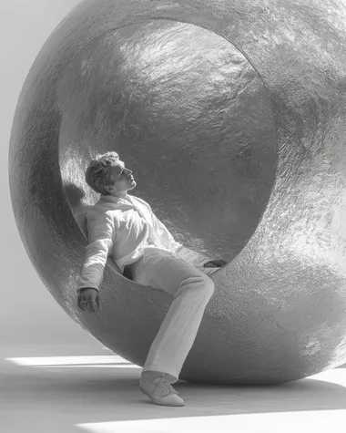 A surreal black-and-white image of a man lying horizontally between two large metallic spheres--one above him, one below. His body is gently pressed between them, as if suspended in stillness. He stretches his arms around the lower sphere, embracing it. The background is pure white, emphasizing the surreal, sculptural composition. The image feels dreamlike, introspective, and artistic, with strong contrast and soft, poetic lighting.   --s 350 --raw
