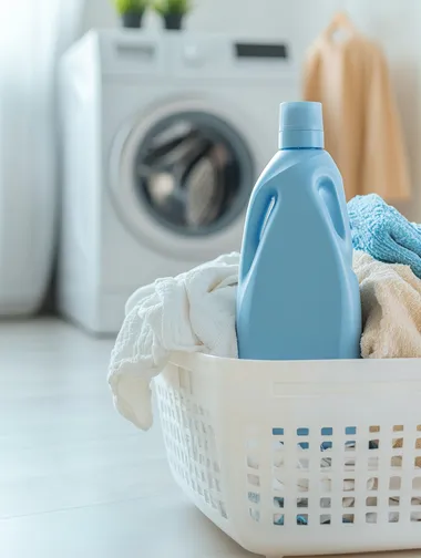 Close-up of a laundry detergent bottle photo of a white plastic basket with a bunch of laundry, laundry in white, blue and beige colors, on the basket there is a large square matte blue bottle of washing gel, feed straight, minimalism, the basket is on the floor next to the washing machine, in the background there is a washing machine, the background is white, atmosphere of cleanliness, laundress banner, white basket.