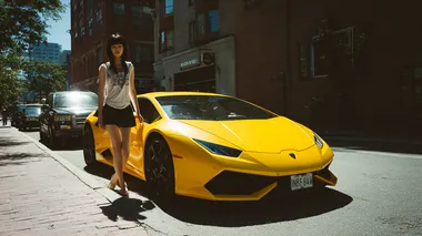 A casual smartphone snapshot of an East Asian woman in a short miniskirt stepping out of a yellow Lamborghini Huracn. Bright daylight, street parking, slightly tilted angle, realistic shadows, no filters.