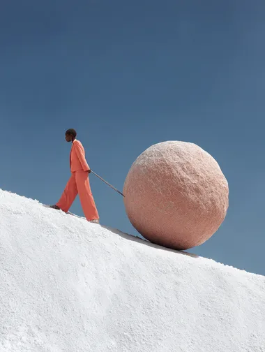 A photograph of a black woman in a salmon-colouted outfit, pushing up a gigantic salmon-coloured boulder up a white chalky hill, in the style of surreal fashion photography representing sisyphus. The image features a blue sky, minimalist design, hyper-realistic rendering, desaturated colours, and high resolution, captured with a Hasselblad camera.  rofile iifez4j