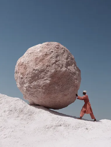 A photograph of a black woman in a salmon-colouted outfit, pushing up a gigantic salmon-coloured boulder up a white chalky hill, in the style of surreal fashion photography representing sisyphus. The image features a blue sky, minimalist design, hyper-realistic rendering, desaturated colours, and high resolution, captured with a Hasselblad camera.  rofile iifez4j