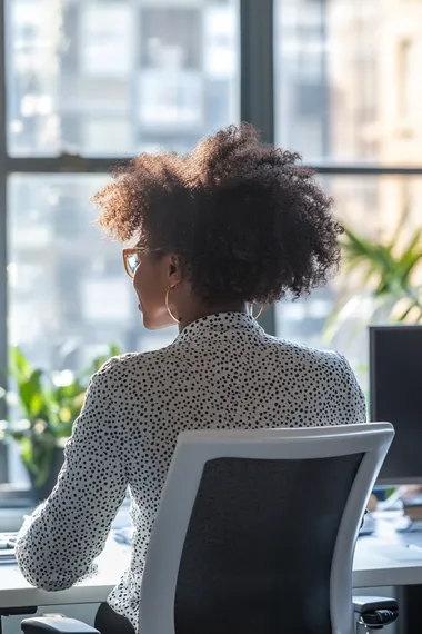 An african female employee sitting at her desk, shot from behind at a slight angle, as she is happily enjoying her work as a project manager, in a well lit, modern office