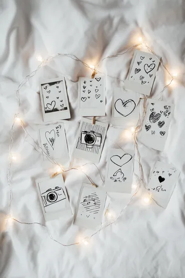 hand-drawn sketch of collection of polaroid photos with doodles and hearts drawn on them, connected by a delicate string of fairy lights, cute,simple, playful strokes, white paper background, soft pencil shading, cozy and charming aesthetic, sketchbook style  --s 50 --raw
