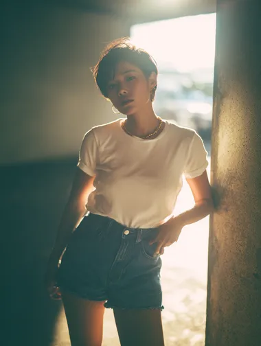A high-end fashion portrait of a short-haired Korean woman, wearing a white T-shirt and denim shorts, standing on a concrete floor, with sunlight shining in from behind. The soft lighting and the contrast of light and dark were captured with a Fujifilm full-frame camera, creating a cinematic bokeh effect