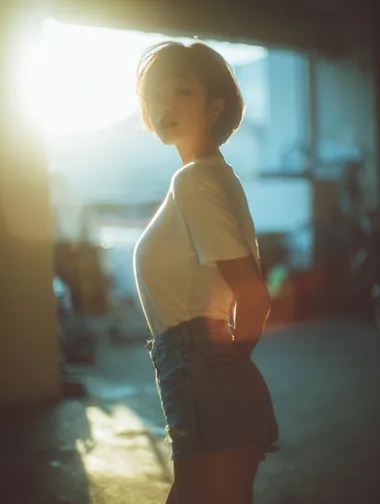 A high-end fashion portrait of a short-haired Korean woman, wearing a white T-shirt and denim shorts, standing on a concrete floor, with sunlight shining in from behind. The soft lighting and the contrast of light and dark were captured with a Fujifilm full-frame camera, creating a cinematic bokeh effect