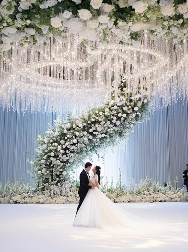 A large screen in front of the stage, a huge crystal spiral chandelier made up of white and green flowers on top with a beautiful wedding couple standing under it, white roses, a large flower wall at ground level, delicate and elegant, a romantic atmosphere, a photographic style, super-realistic, high-resolution, professional lighting, and insanely detailed.