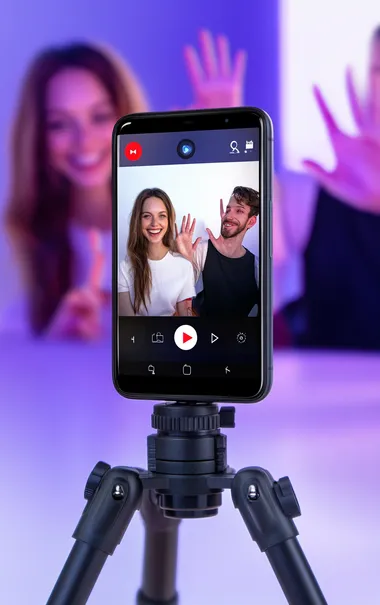 A smartphone with an open camera app, showing two people in the background smiling and waving at each other on a video call. The person to be used as an AI model is facing forward towards us, wearing black pants and a white shirt. On top of that, we see a red recording button. The background color is a purple-blue gradient. In front of them, there is also a visible part of a tripod holding the phone.