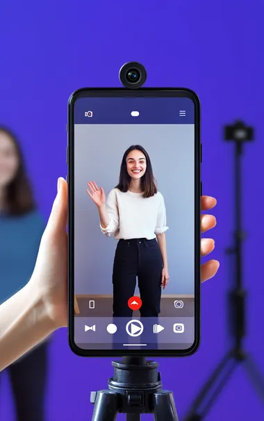 A smartphone with an open camera app, showing two people in the background smiling and waving at each other on a video call. The person to be used as an AI model is facing forward towards us, wearing black pants and a white shirt. On top of that, we see a red recording button. The background color is a purple-blue gradient. In front of them, there is also a visible part of a tripod holding the phone.