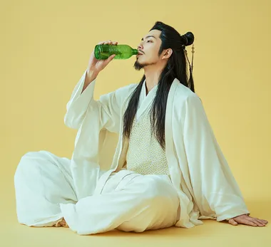 A man in ancient Chinese costume, wearing black hair and a small beard, is sitting on the ground with his legs crossed, drinking from a green plastic bottle. He is wearing a white long-sleeve robe with a light yellow dot-patterned shirt. His mouth is slightly open, and the image is a full-body shot against a solid color background, with studio photography, natural lighting, medium distance, and a clean, cool-colored background.