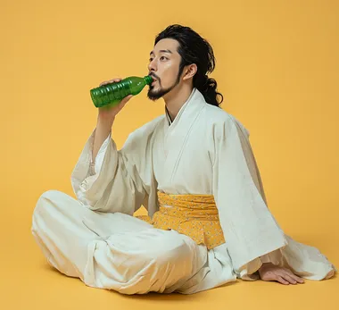 A man in ancient Chinese costume, wearing black hair and a small beard, is sitting on the ground with his legs crossed, drinking from a green plastic bottle. He is wearing a white long-sleeve robe with a light yellow dot-patterned shirt. His mouth is slightly open, and the image is a full-body shot against a solid color background, with studio photography, natural lighting, medium distance, and a clean, cool-colored background.