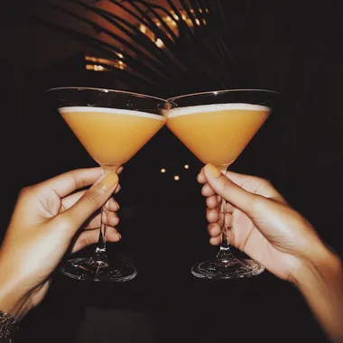 two hands holding two frothy orange martini's must look like a club like photo, high energy and realistic