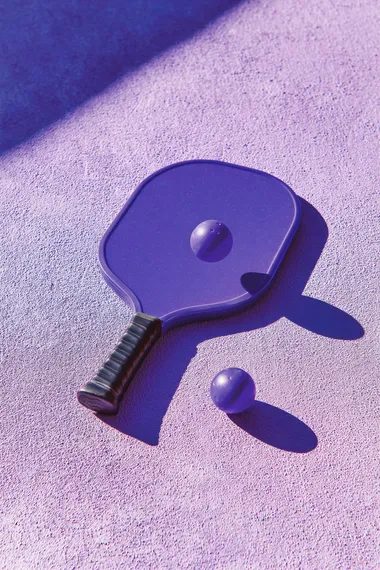 trendy pickleball paddle with ball on textured lavender surface, high angle summer sunlight casting bold shadow, minimal composition, no text