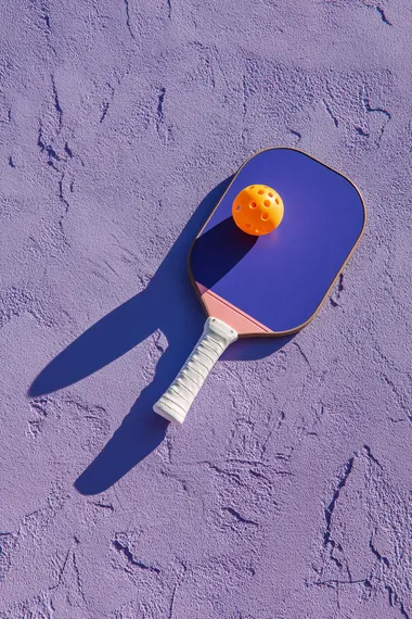 trendy pickleball paddle with ball on textured lavender surface, high angle summer sunlight casting bold shadow, minimal composition, no text